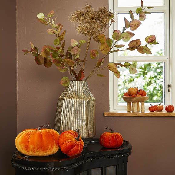 efterårsdekoration med kunstige efterårsgrene i banket metalvase og velourgræskar sat på et lille bord ved vindue med små kunstig græskar i en lille træskål
