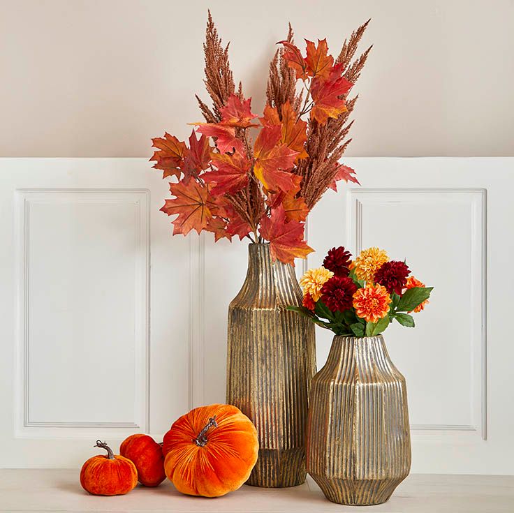 Efterårsopsætning med kunstige dahlia blomster, bladgrene og pampas i vaser af banket metal, små velourgræskar i to størrelser langt ved siden af.