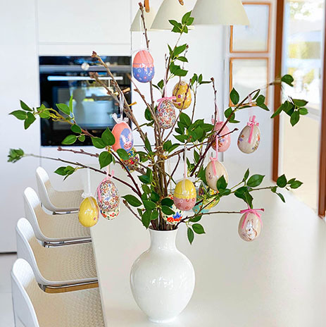 Kunstige grene med grønne blade pyntet med farverige påskeæg af papir på køkkenbord