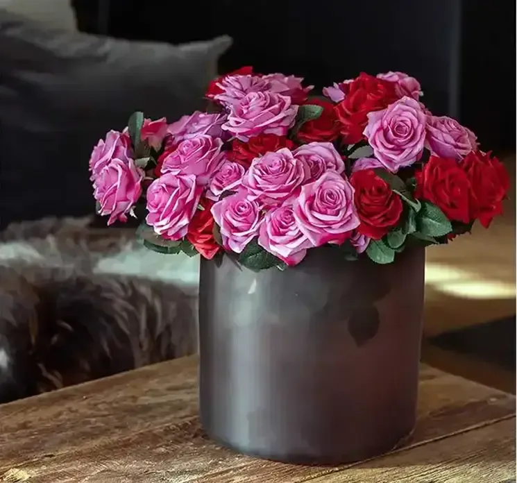 Vase af glas i blå nuancer vist på stuebord med kunstige roser i pink og røde