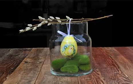 Påskeæg der hænger ned i en klar glasvase på en kunstig gæslingegren, og med kunstige mossten i bunden af vasen