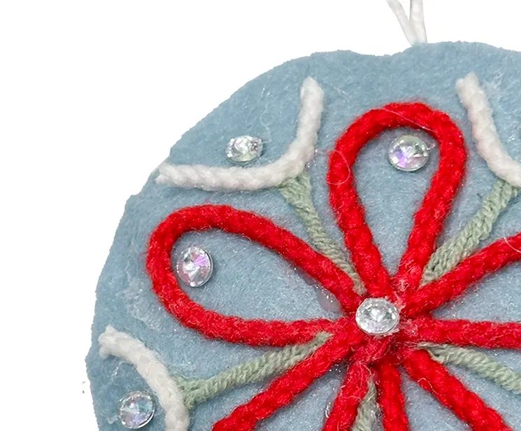 Filt kugle i blå med pynt på en side, kuglen er flad og med snor til at hænge op closeup