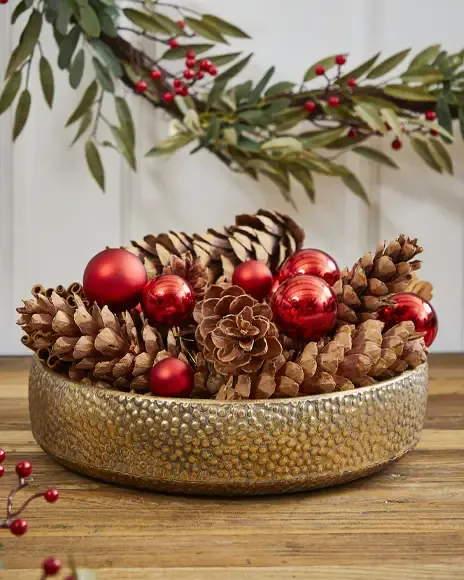 Fin juledekoration i guld skål fyldt med grankogler og små røde plast julekugler 