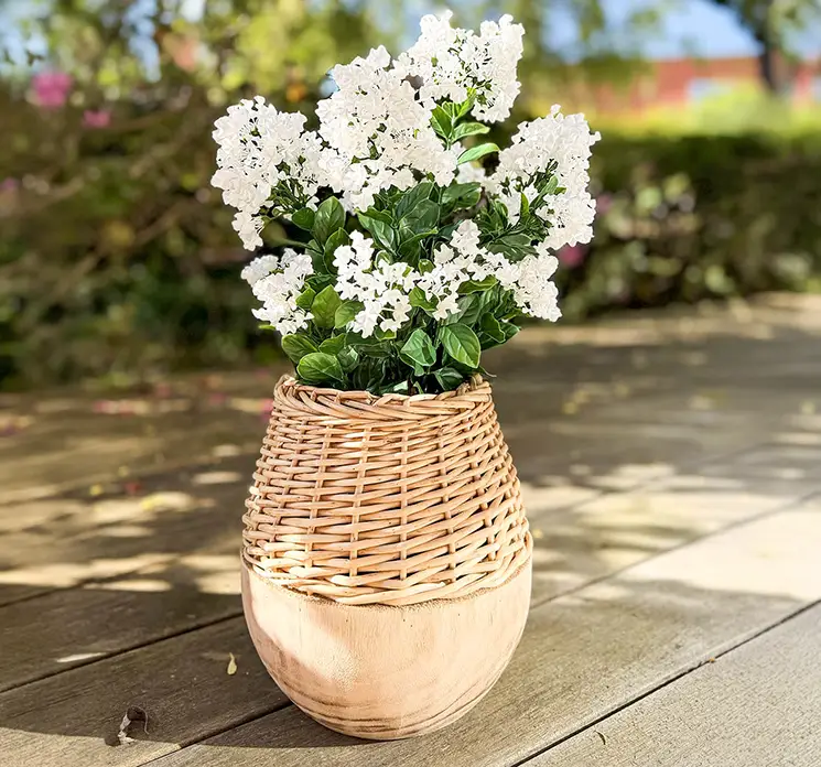Syren i hvid kunstig blomst vist i krukke af paulownia træ og flet i havemiljø