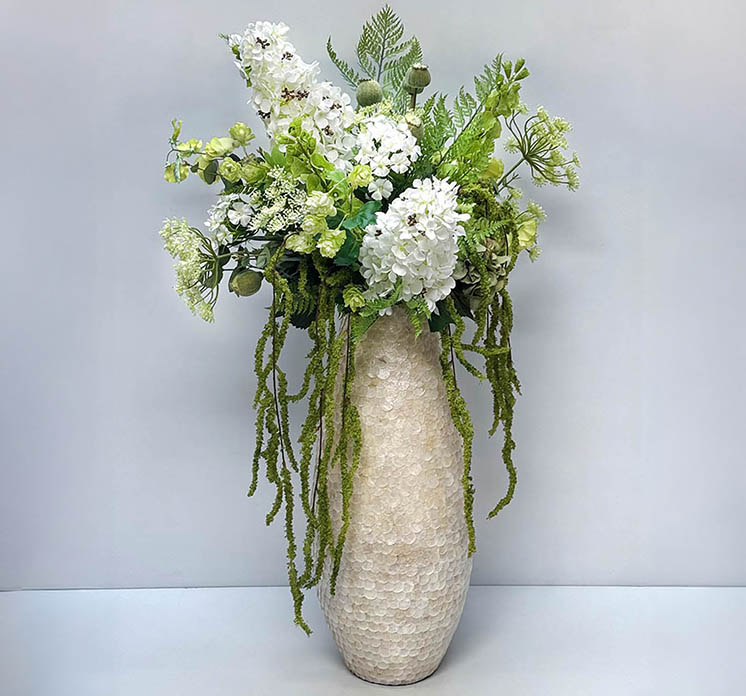 Buket sammensat af kunstige blomster og grene i hvide og grønne sat i bambus vase med skæl af muslinger i creme farvet