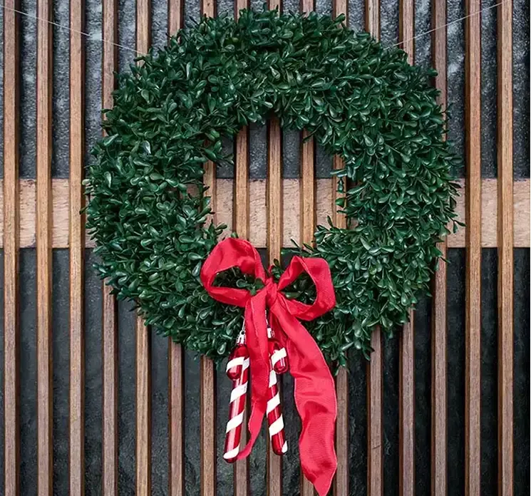 Kunstig buksbom krans i grøn plast uv beskyttet vist hængende på dør med rødsløjfe og slikstokke af plast