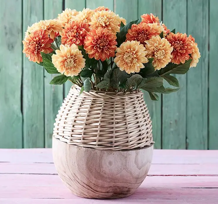 Dahliaer af plast i orange og gule sat sammen i en trævase med flet, kunstig blomst
