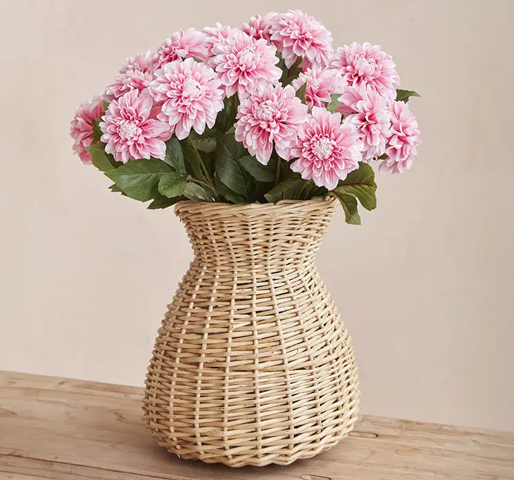 Vase formet som kande af flettet rattan i naturfarvet fyldt med flotte kunstige dahlia i lyserøde