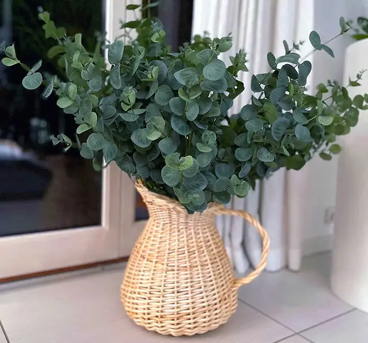 Vase formet som kande af flettet rattan i naturfarvet fyldt med kunstige eukalyptus grene vist i køkkenmiljø