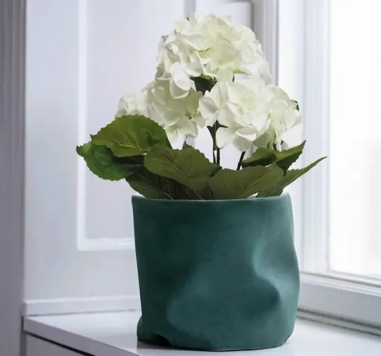 Potteskjule i grøn af læder vist med en kunstig pelargonie i vindueskarm