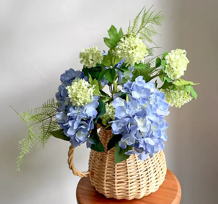 Hortensia i blå og kalkved i grøn kunstige blomster sat sammen i en smuk buket i flettet vase kande