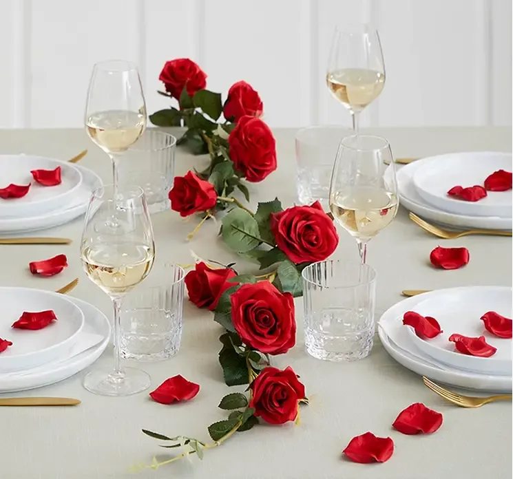 Kunstige rosenblade af satin i rød vist på et dækket bord med kunstig rød rosenranke