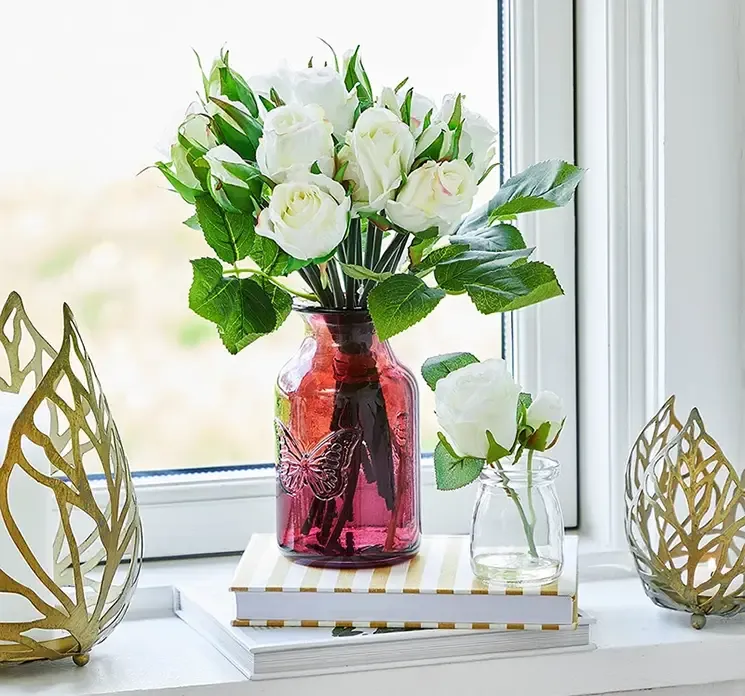 Vase af glas i mørk pink med sommerfugle motiv vist i vindueskarm med kunstig rosenbuket i hvid
