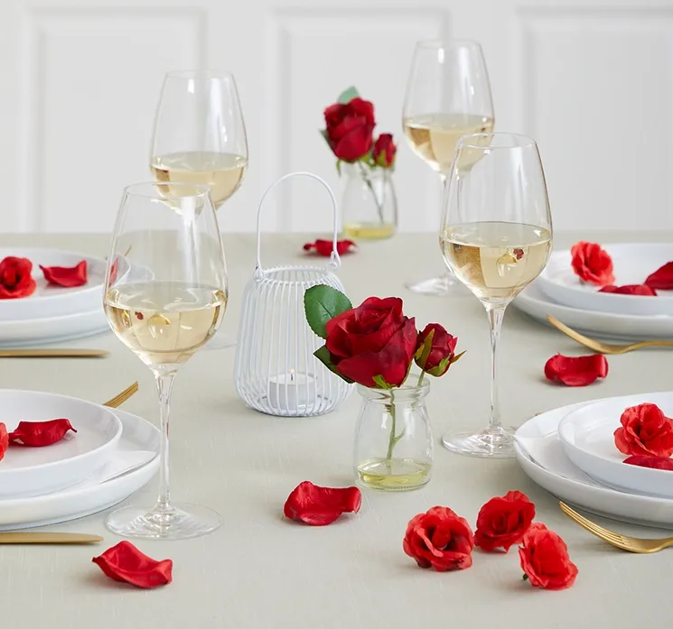 Bord dækket med kunstig rød rose i glasvase, små rosenhoveder og rosenblade spredt ud på bordet.