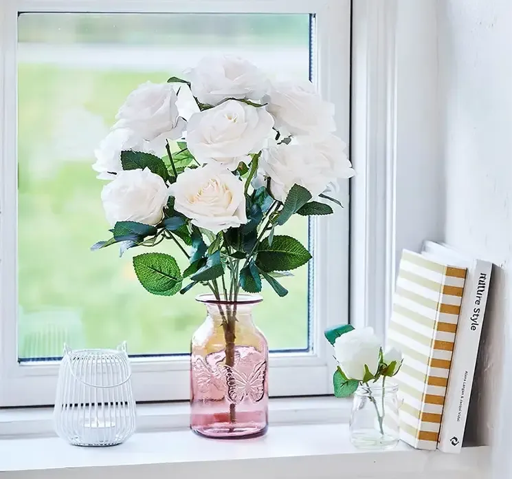 Vase i lyserød af glas med sommerfugl vist med en kunstig buket af hvide roser i en vindueskarm