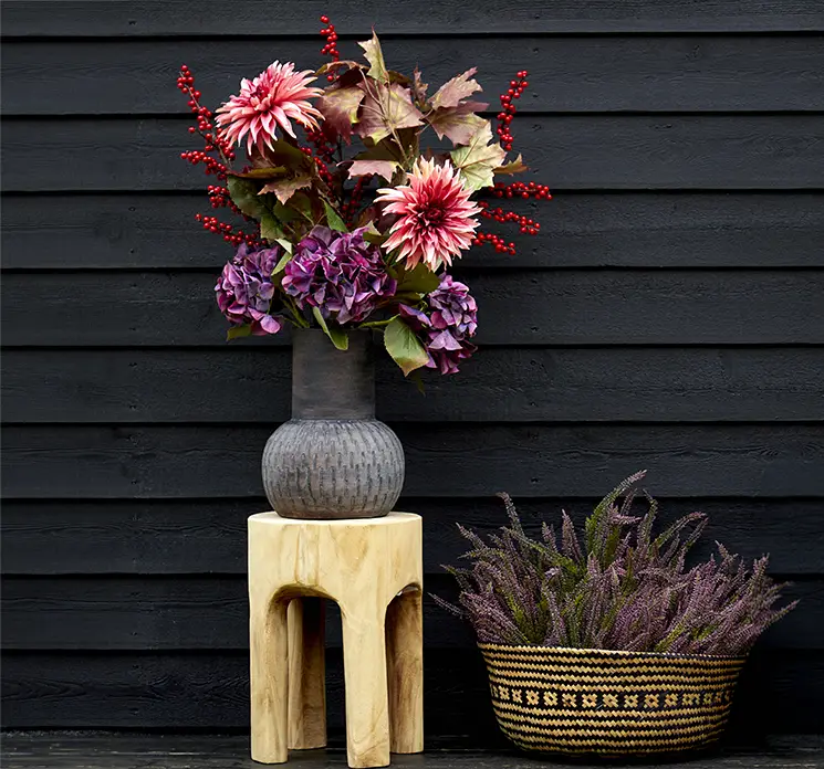 Vase af terracotta vist på terrasse med kunstig buket af blandt andet dahlia og fletkurv ved siden af med kunstige lyngbuketter