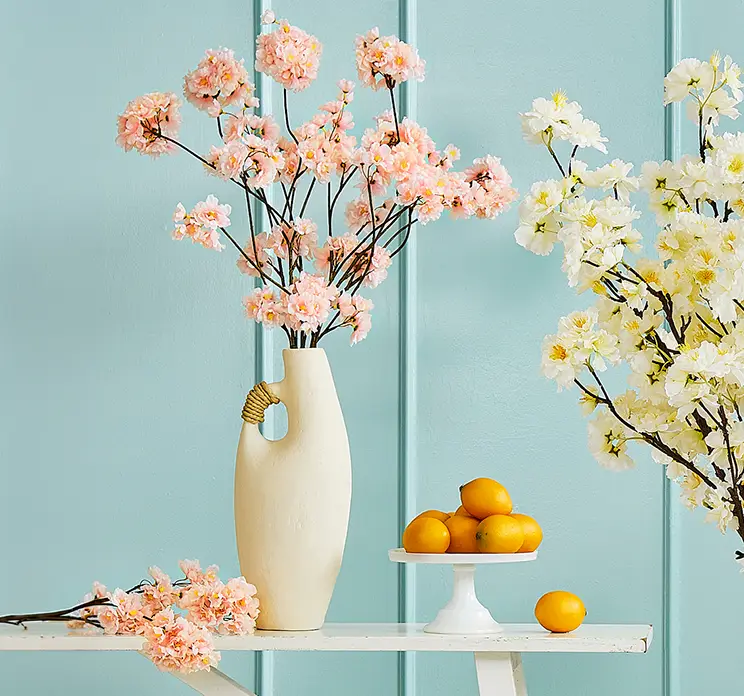 Kirsebærgren af plast kunstig gren med små ferskenfarvede blomster sat i en lys vase af papmache i stuemiljø