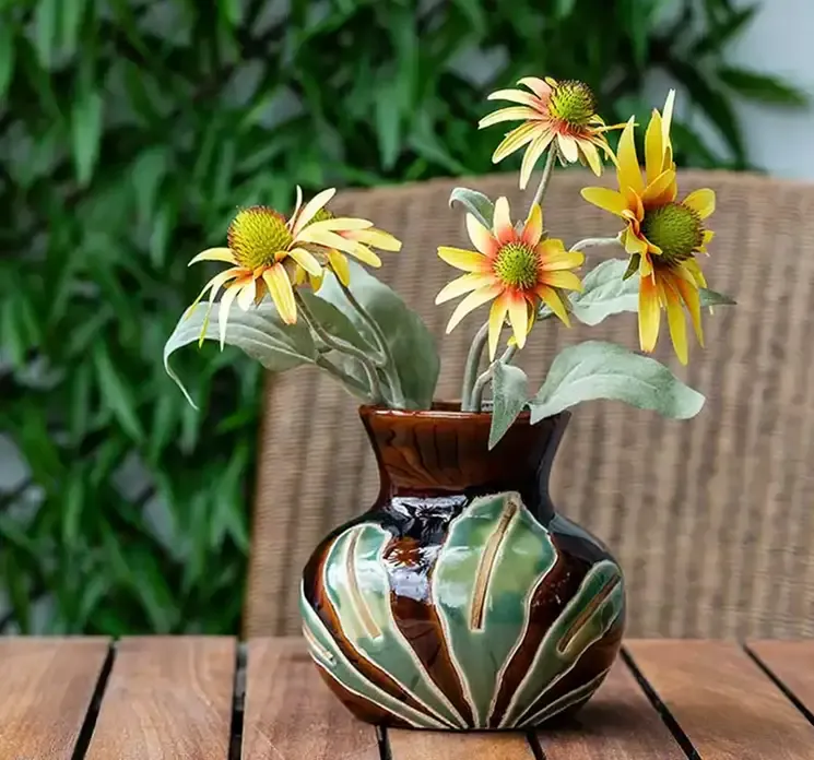 Håndlavet vase af porcelæn med blad motiv, vasen er brun og grøn vist på et havebord med kunstige gule solhatte