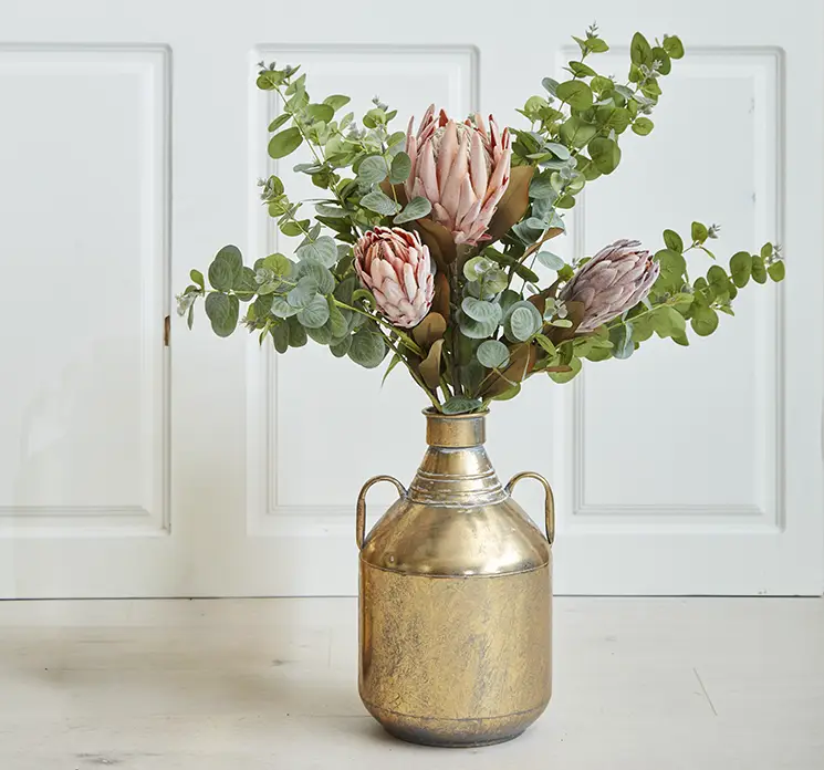 Vase af metal i guld look med hanke vist med kunstige protea blomster og eukalyptus grene